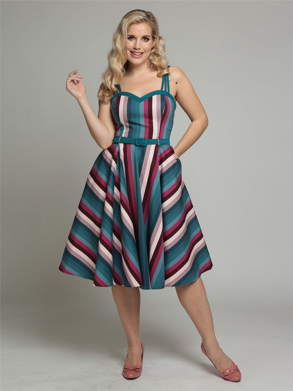 blonde vintage pin up model wearing a turquoise, pink, white and burgundy asymmetrical striped swing dress and pink pumps