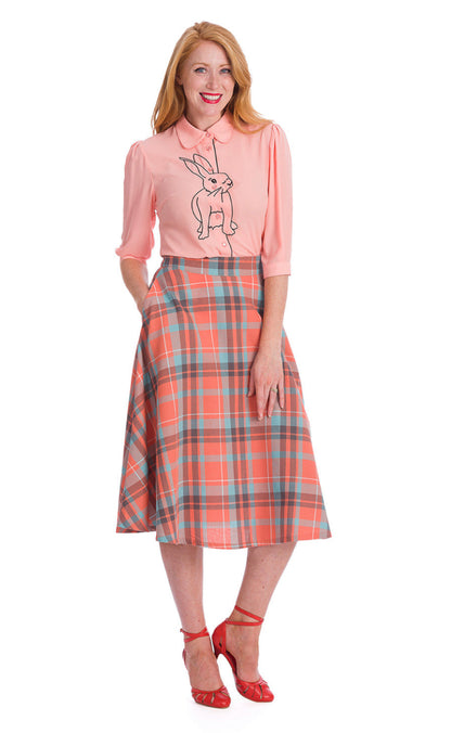 Smiling women standing with one hand in her skirt pocket. She is wearing a light orange and blue check skirt that falls just below the knee and a light pink blouse with a black embroidered rabbit on the front.