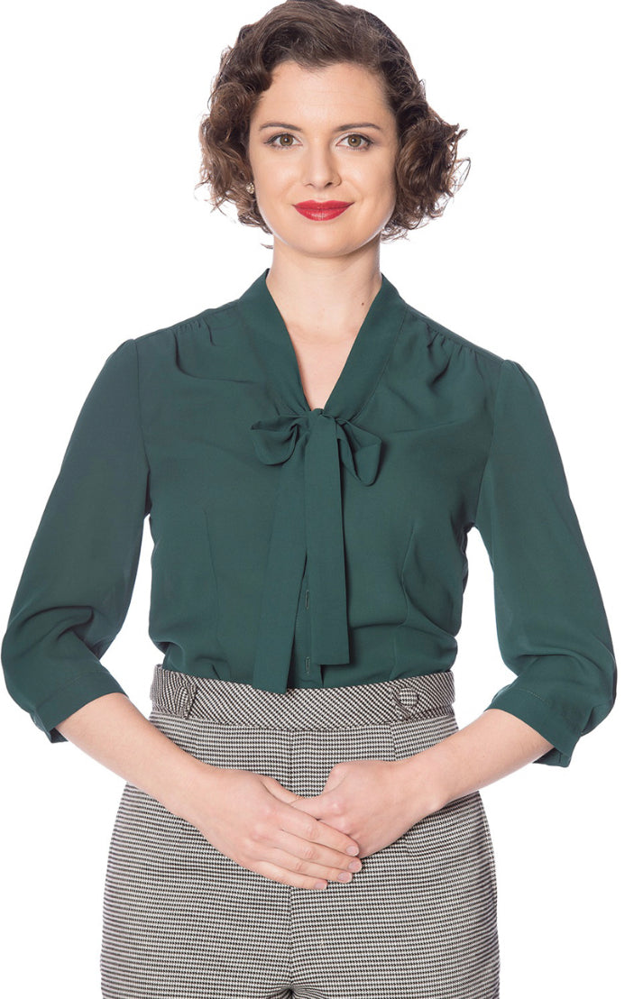 Smiling woman wearing red lipstick, a green 40s style pussybow tie neck blouse with 3/4 length sleeves and a button up front tucked into houndstooth high waisted trousers.