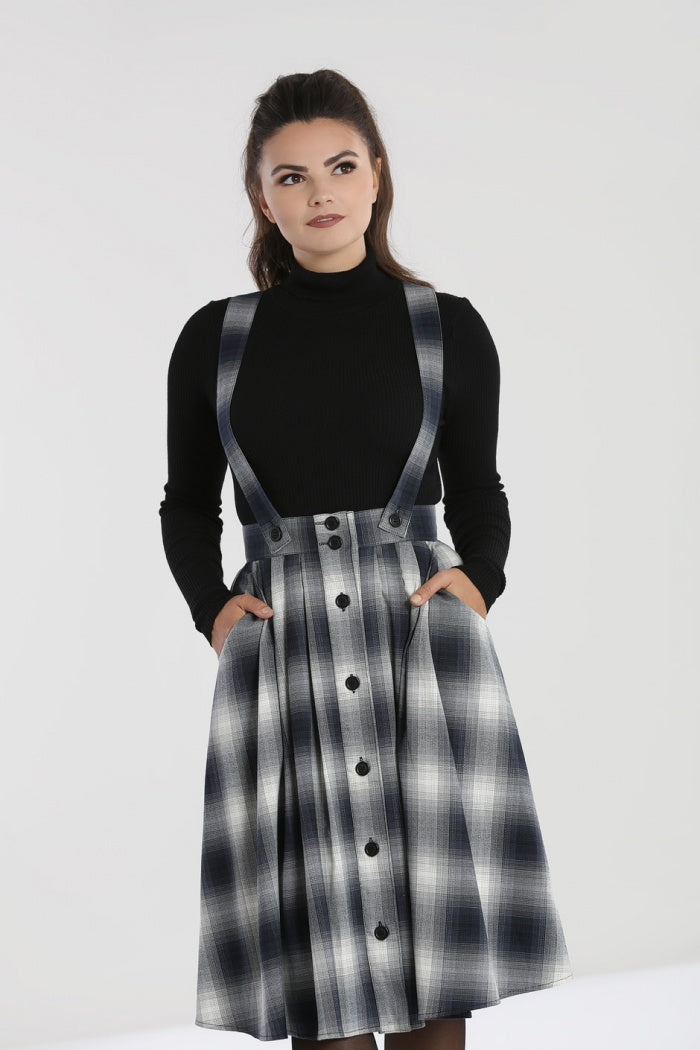 Brunetter woman standing with both hands in her pinafore skirts front pockets. She wears a black turtleneck sweater underneath.