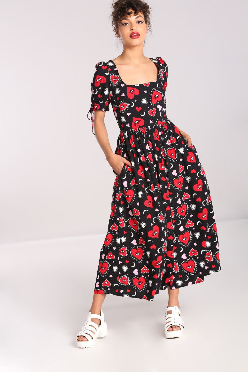 Tall slim woman with brown curly hair tied back standing in a black maxi dress with an all over red heart print and white sandals