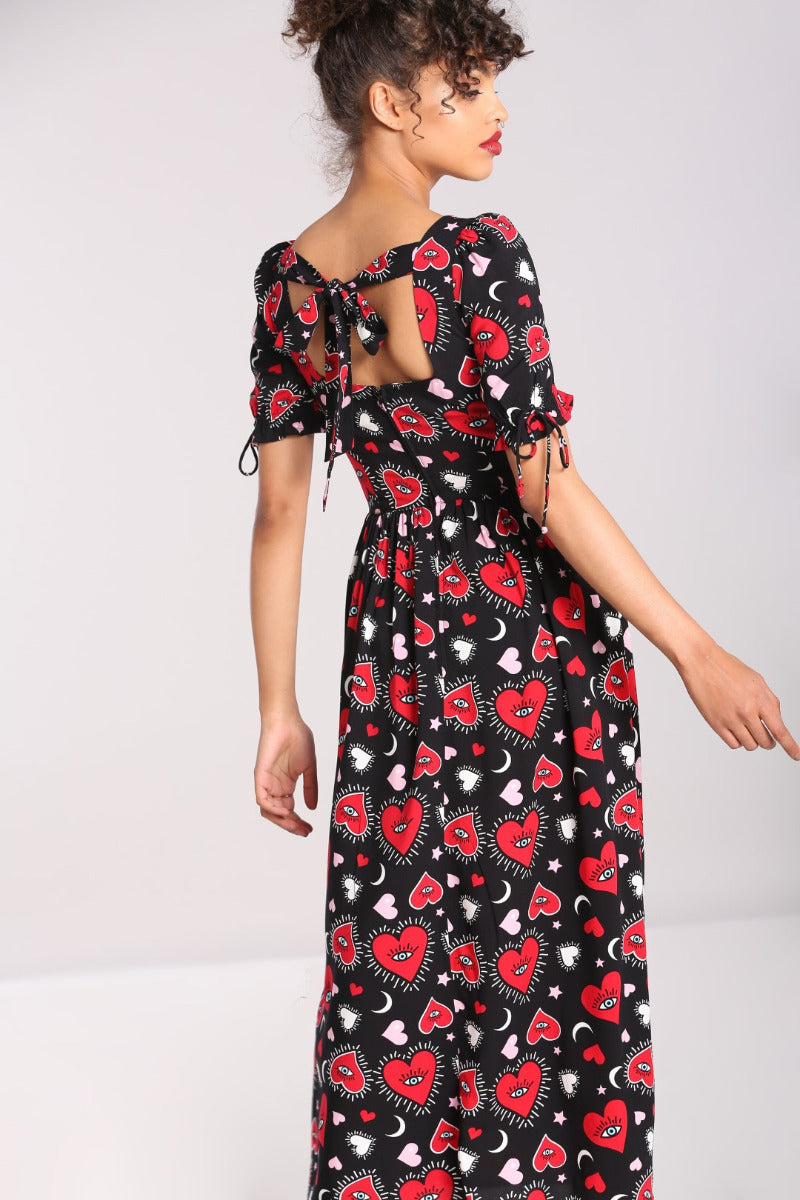 Elegant woman with her curly brown hair tied back in a bun showing the back of the Kate Heart Maxi Dress which features a pretty tie detail at the back by her neck and a square neckline, short sleeves and a semi fitted waist.