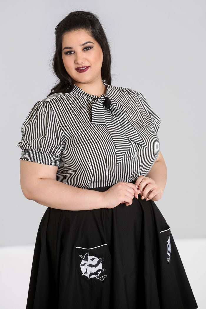 Smiling woman wearing dark red lipstick standing holding her hands together in front of her waist. She is wearing the Hell Bunny Humbug blouse in black and white and a black skirt with a white moon and flying bat motif on the front pockets. 