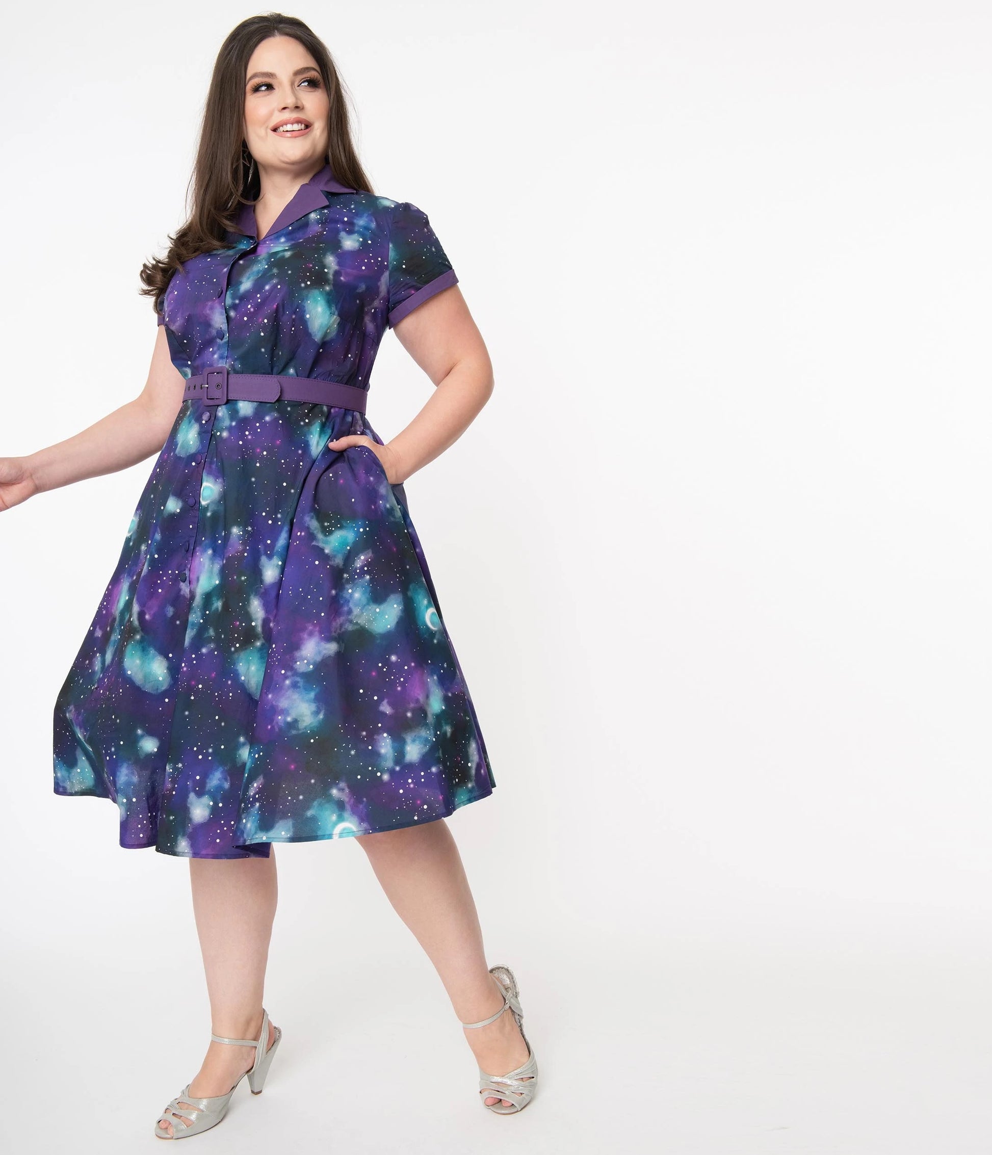 A brown haired smiling girl wearing a purple space print dress standing looking over her shoulder in front of a white background