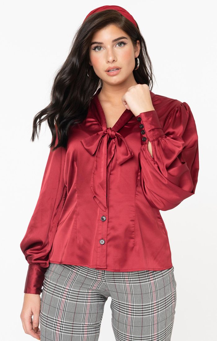 Pretty brown haired girl in natural makeup with one hand up near her face stands wearing an untucked burgundy blouse and grey checked skinny trousers