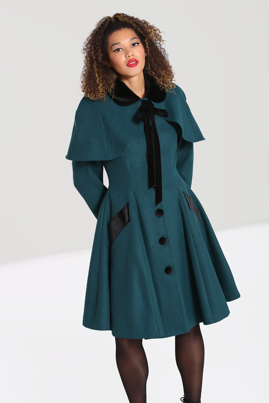 Glamorous woman with curly brown hair and 50s makeup wearing a dark green vintage style swing coat and a shoulder cape tied at the neck with a velvet bow
