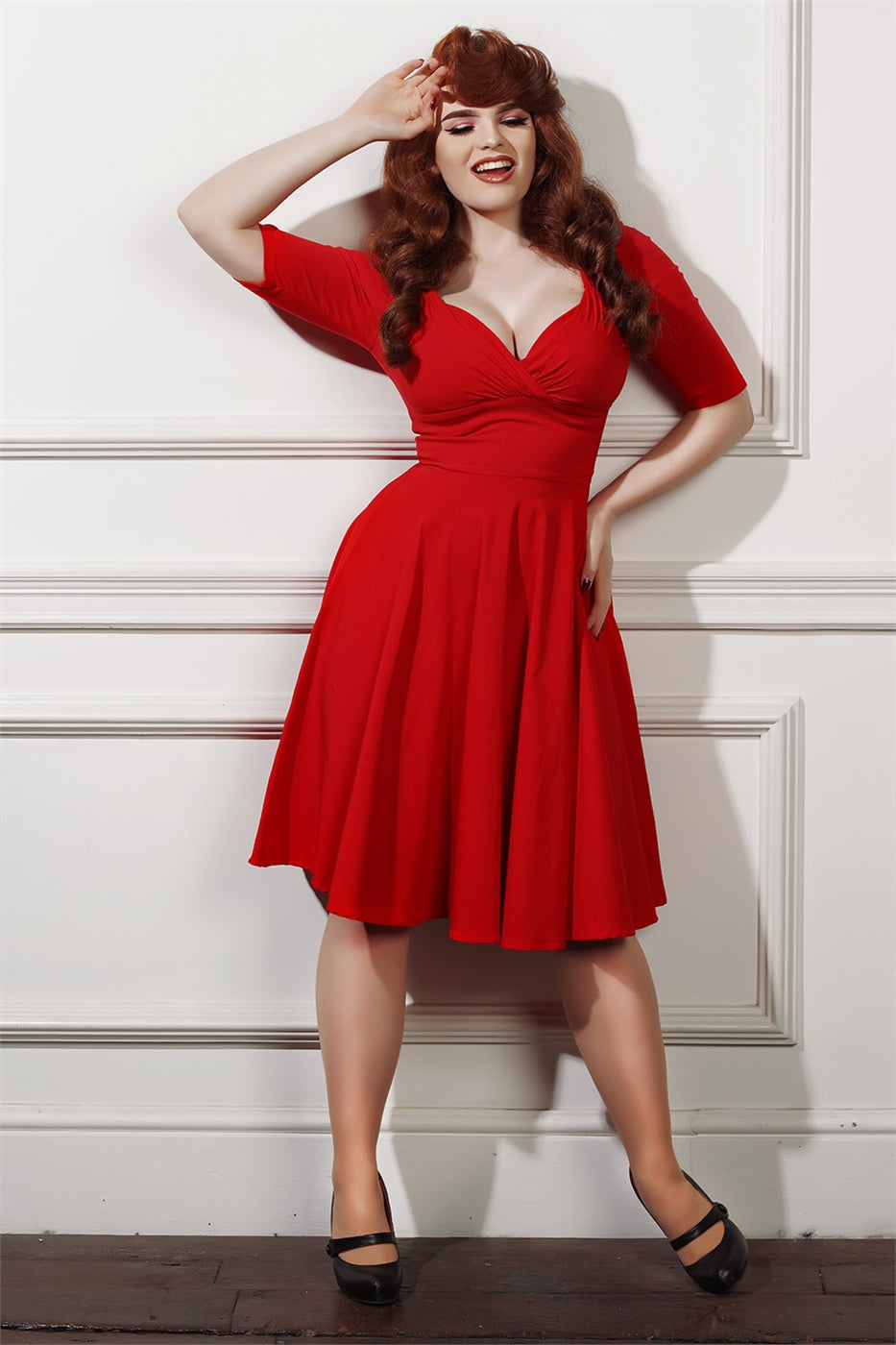 fair skinned model smiling with her hand up to her forehead and long curly auburn hair wearing a red swing dress standing in front of a white wooden panelled wall.