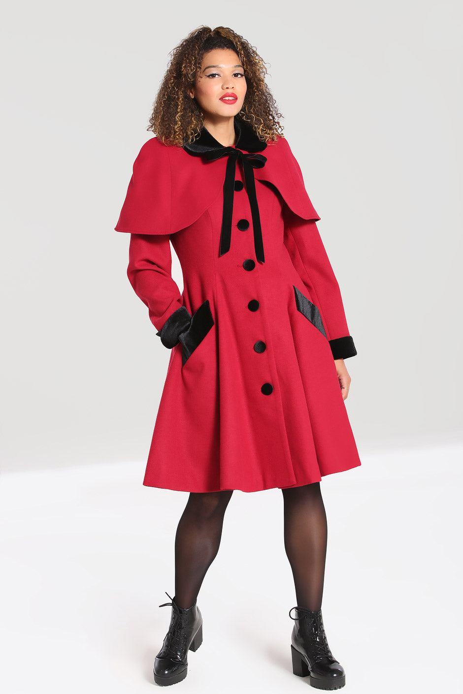 brunette curly haired smiling woman wearing a long red coat standing with one hand in her coat pocket 