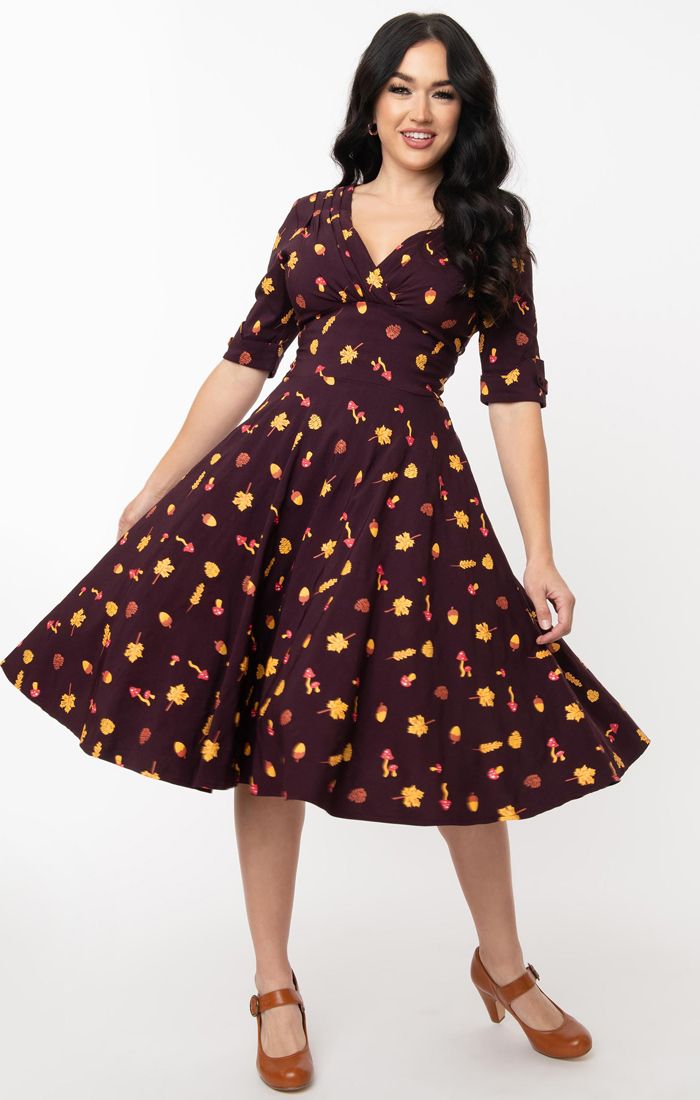 Smiling, glamorous girl wearing  deep purple 50s inspired fit and flare dress with an allover print of autumn leaves, mushrooms and acorns