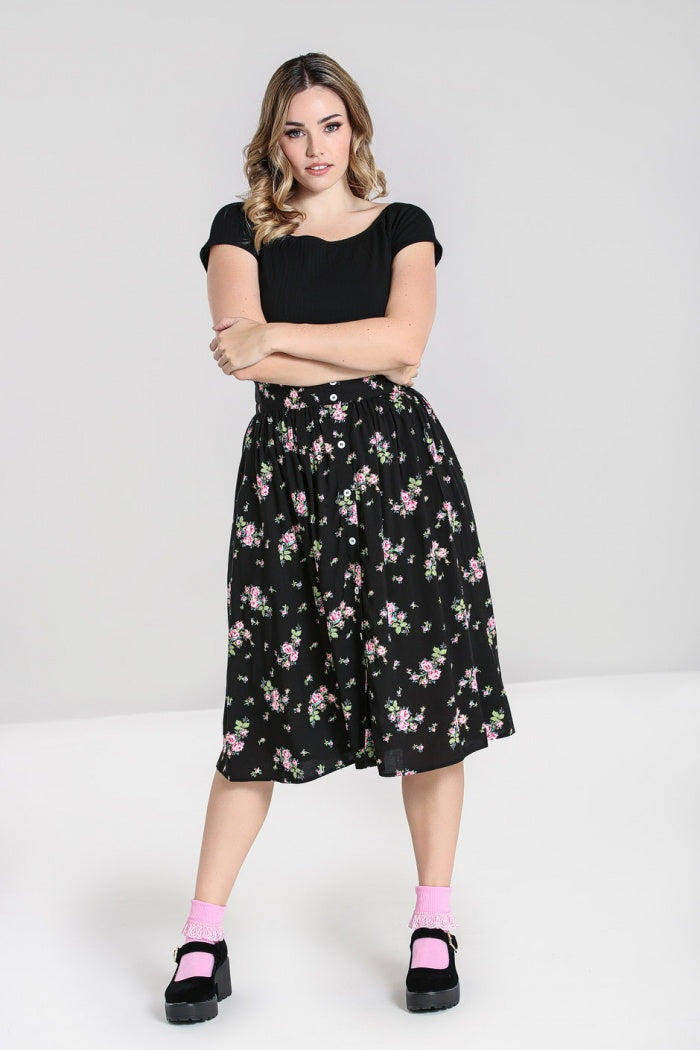 Blonde haired girl with blue eyes and natural makeup standing with her arms folded  wearing a black top, floral skirt and pink socks with black shoes