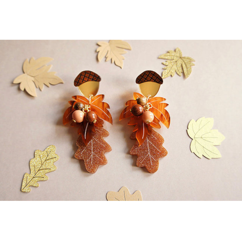Gold and brown paper leaves scattered around to frame a pair of handmade Autumn leaf design earrings with acorns and wooden balls in the centre that resemble berries
