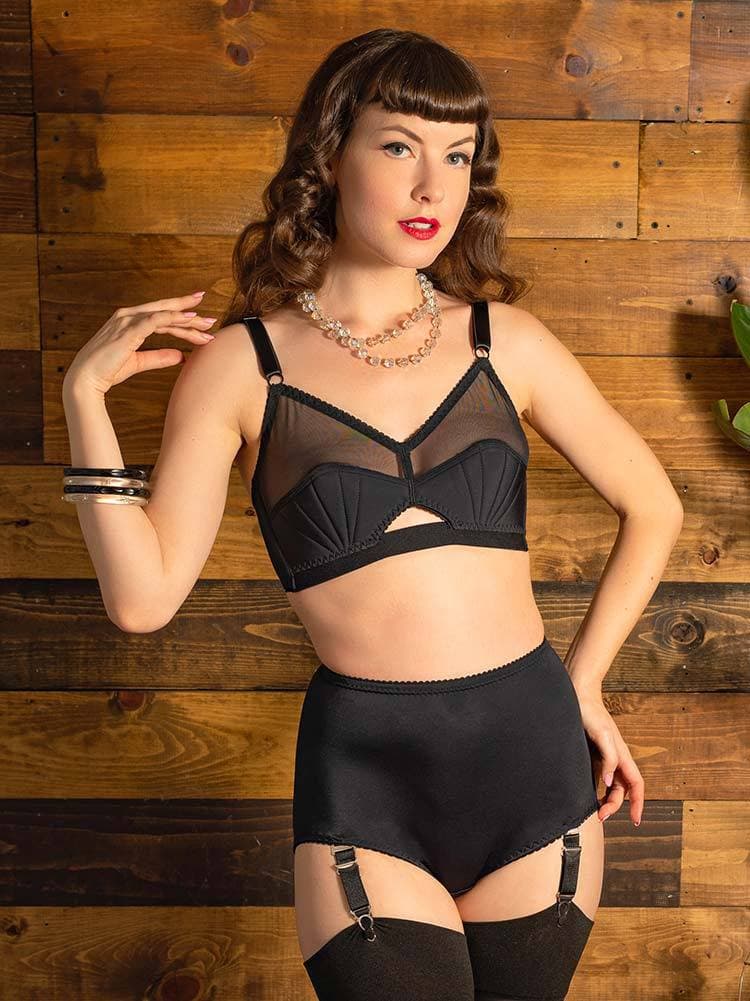 Glamorous woman with brown curled hair standing with one hand on her hip and the other touching her shoulder. She is wearing the Liz high waisted black suspender knickers  and matching bra and black stockings