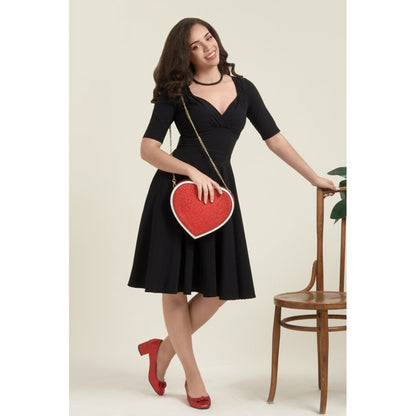Smiling model wearing a red heart shaped handbag, black dress and red stilettos leaning against a chair