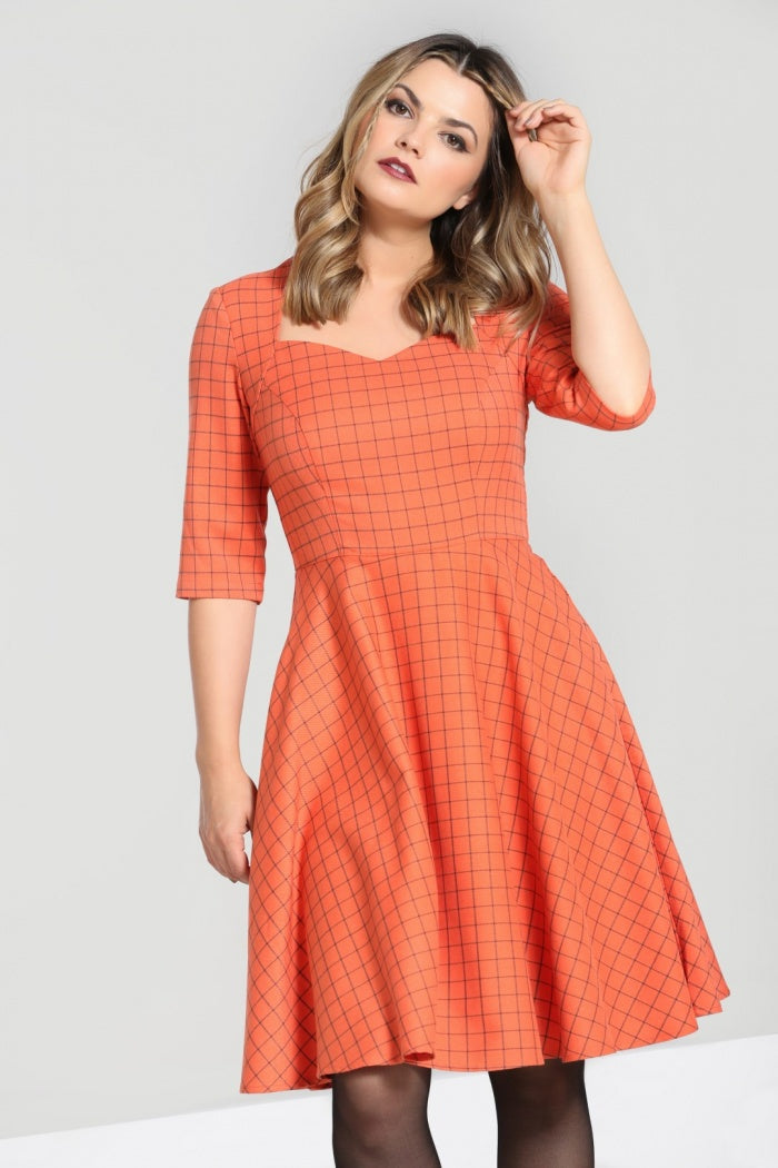 Blonde model wearing natural makeup plays with her hair while wearing a sweetheart neckline mid length orange dress with fine black lines in a checked pattern.