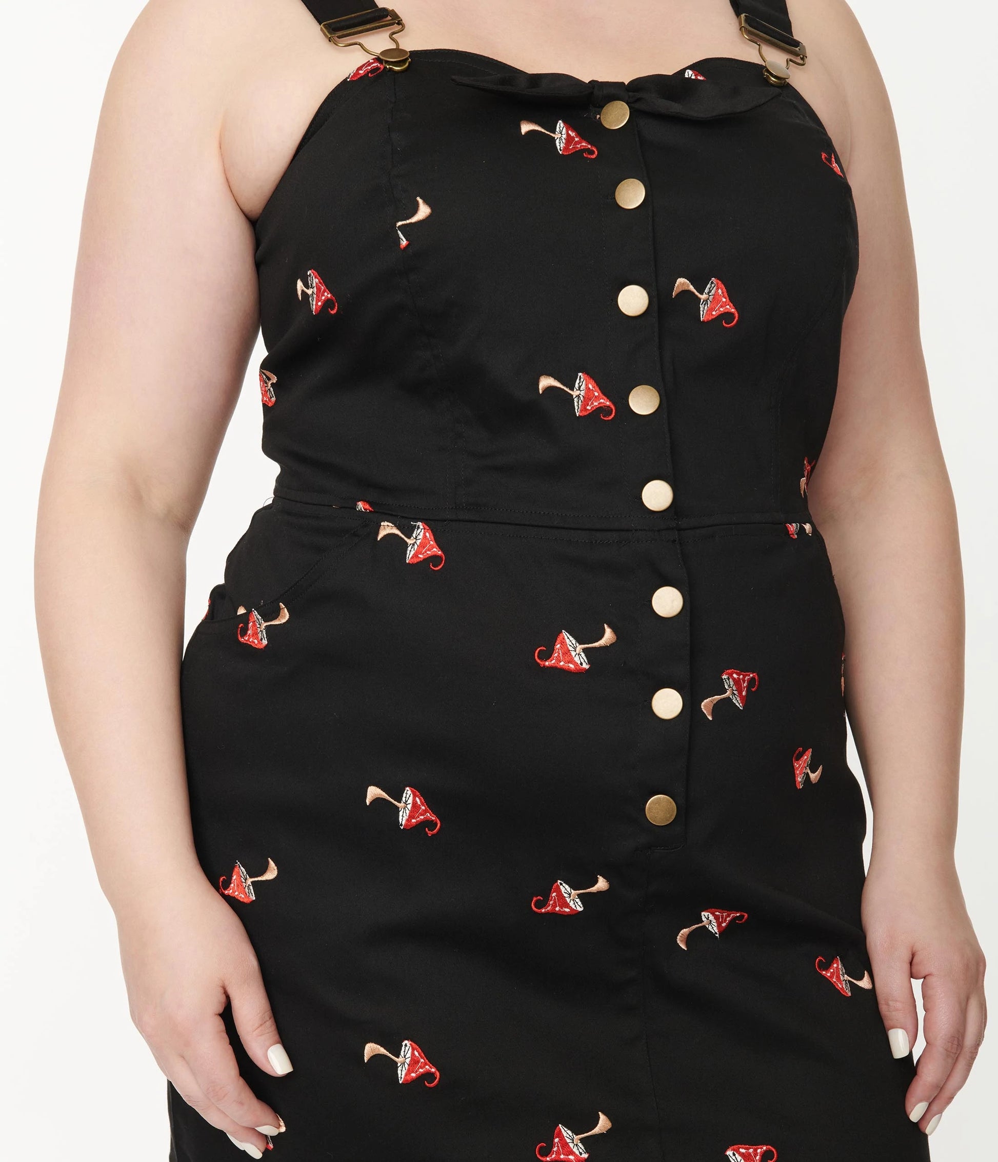 Close up of the button front black denim pinafore dress featuring embroidered red and white mushrooms being worn by a plus size model and a plain white background behind.