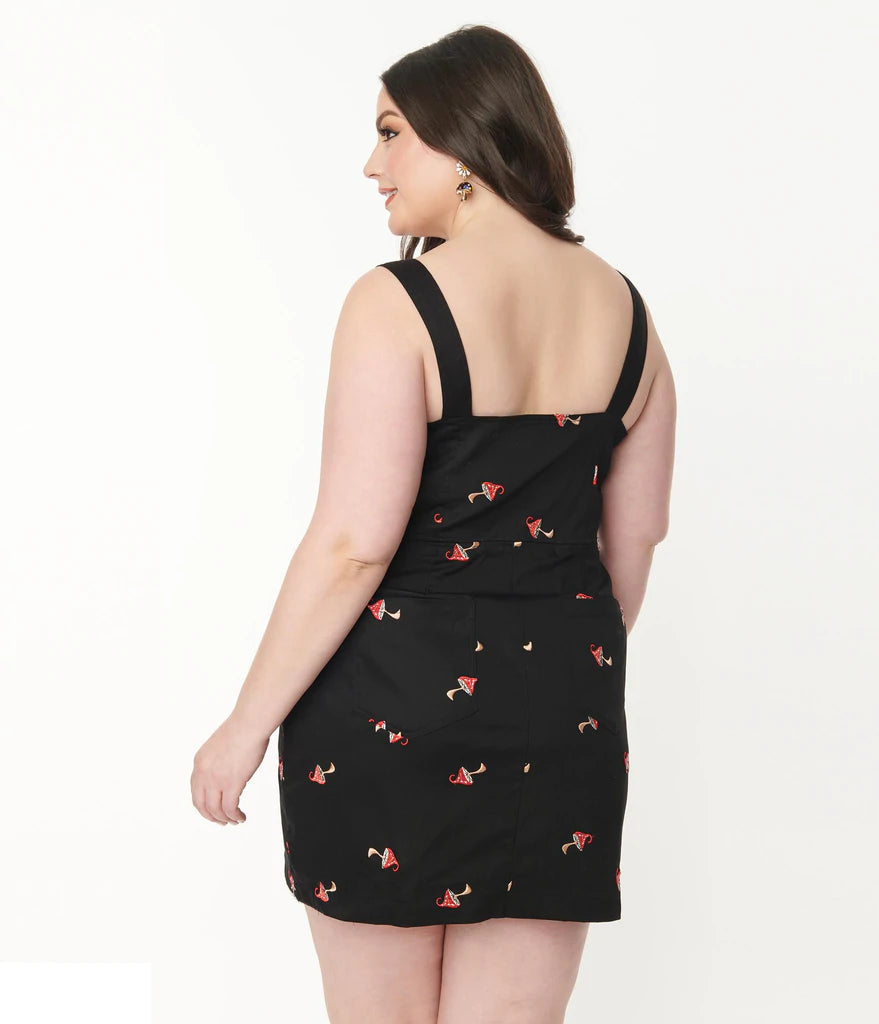 Dark haired smiling plus size girl standing facing away showing the back of the black denim pinafore dress with embroidered red and white mushrooms.