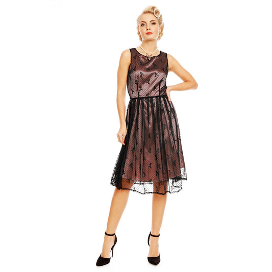 Blonde model with short curly hair stands with one hand on her hip wearing a dark pink satin dress with a black mesh overlay 