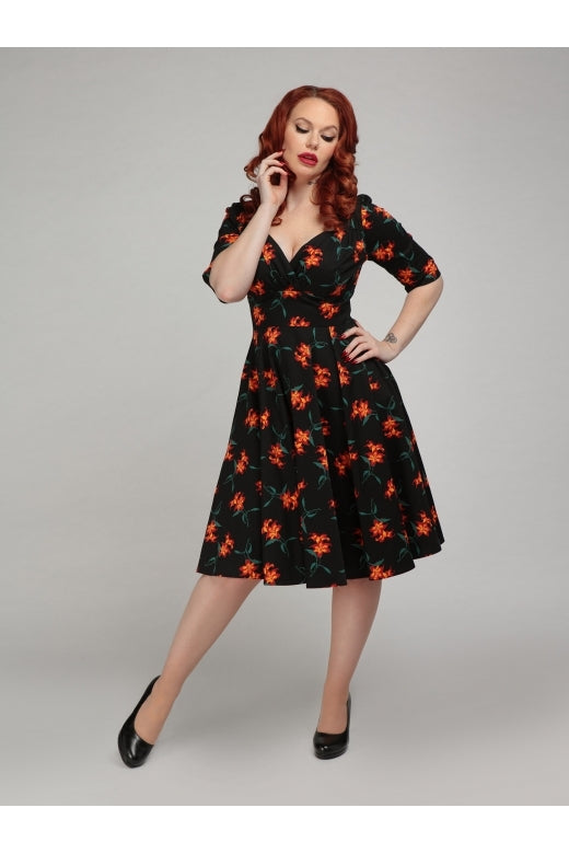 A ginger haired vintage styled woman standing with one hand on her hip wearing high heels and a black fit and flare dress with orange flower pattern