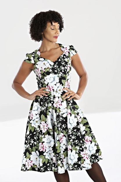 Elegant woman with dark curly hair standing with her hands on her hips. She is wearing a mid length 50s style fit and flare dress with a white and pink floral pattern all over a black background