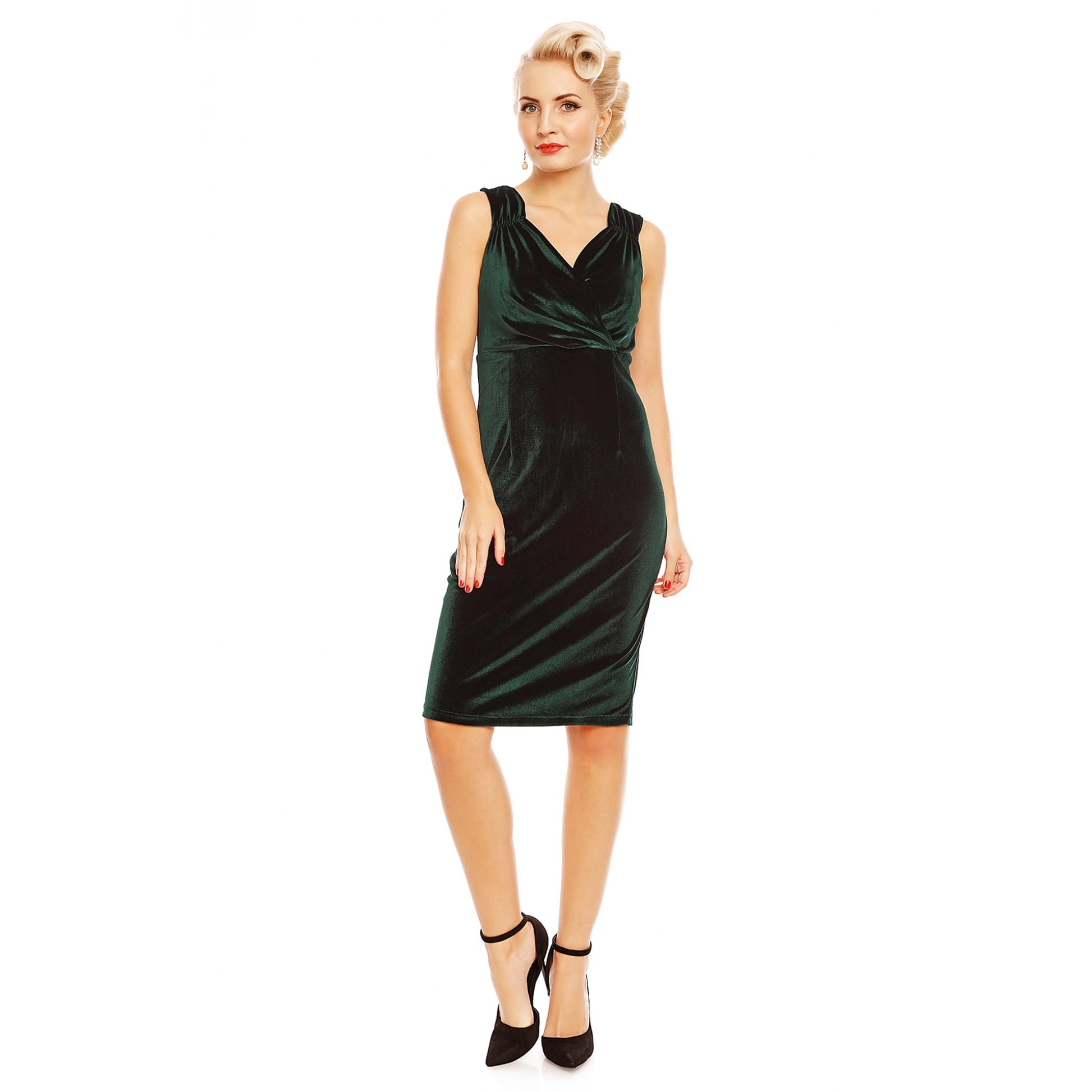 Sultry model with vintage styled makeup and short blonde victory rolls and curled hair stood wearing a classy sleeveless green velvet mid length dress and strappy black high heels