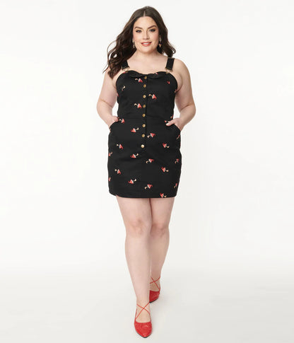 Plus size dark haired smiling girl standing with hands in pockets of black strappy denim pinafore dress with embroidered red and white mushrooms and red strappy shoes