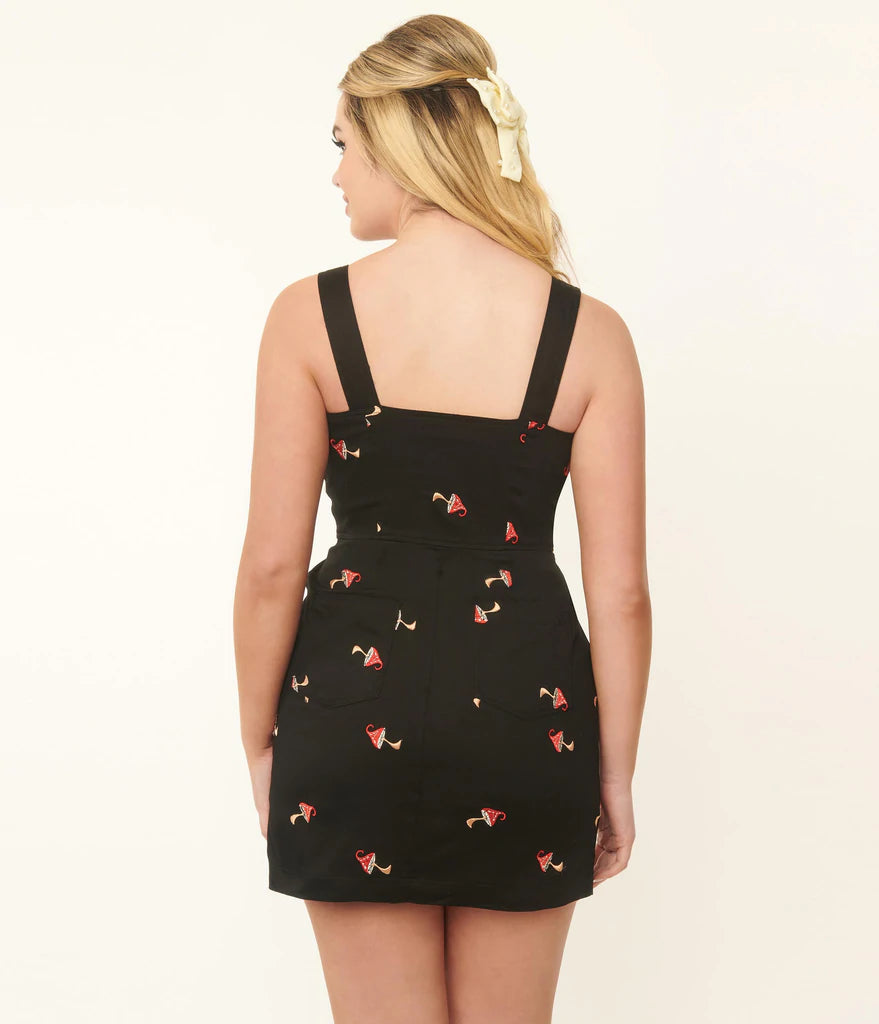 Back view of blonde girl wearing a strappy black denim pinafore dress with embroidered red and white mushrooms all over