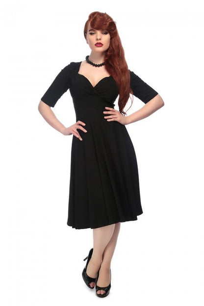 Sultry long haired model wearing red lipstick, a black mid length 50s style dress and black stilettos