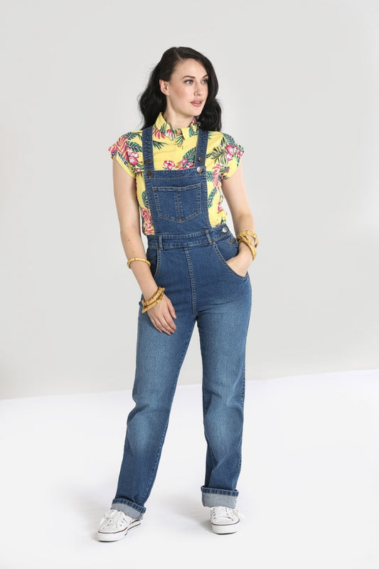 Brown haired girl wearing a floral pattern shirt and blue denim dungarees with white trainers.
