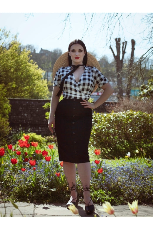 Elegant dark haired woman wearing a black and white gingham wrap top and Bettina pencil skit standing in a country garden