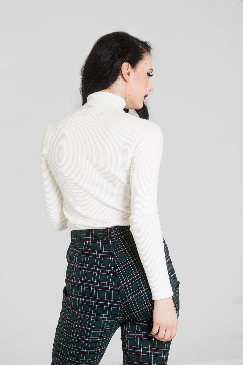 woman with black hair standing with one arm by her side facing away from the camera. She is wearing the  ivory Spiros top by Hell Bunny and green tartan high waisted trousers.