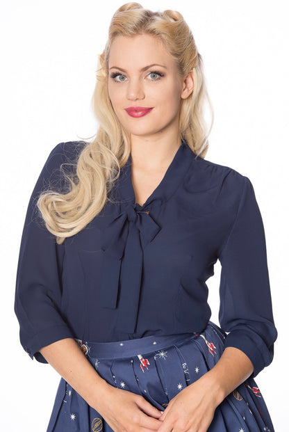 blonde woman holding her hands in front of her wearing a navy 40s style pussybow tie neck blouse with 3/4 length sleeves.