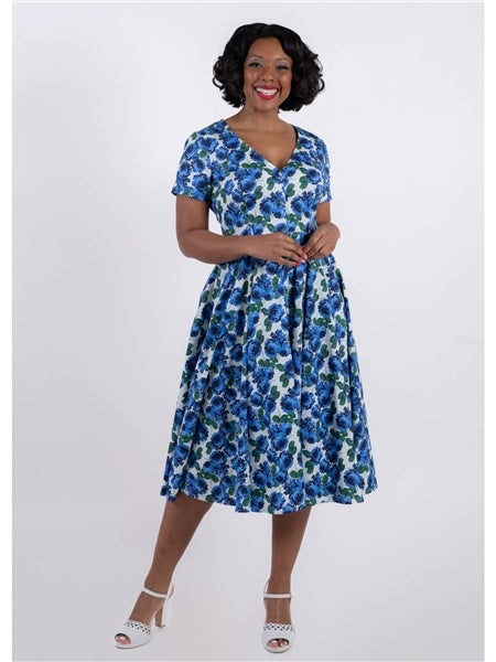 A woman standing, wearing a blue and green floral swing dress with a V-neckline and short sleeves. She is also wearing white sandals and has a belt, though the belt is not included in the image.