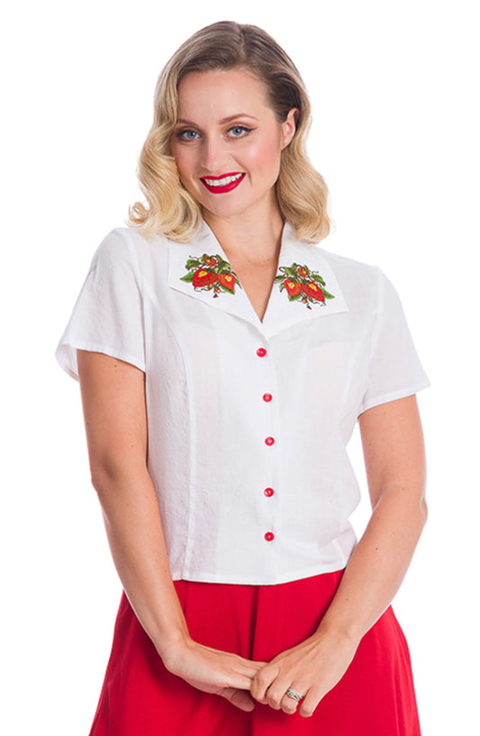 Smiling woman wearing red lipstick, a white blouse with red buttons and embroidered strawberries on the collar and red trousers.