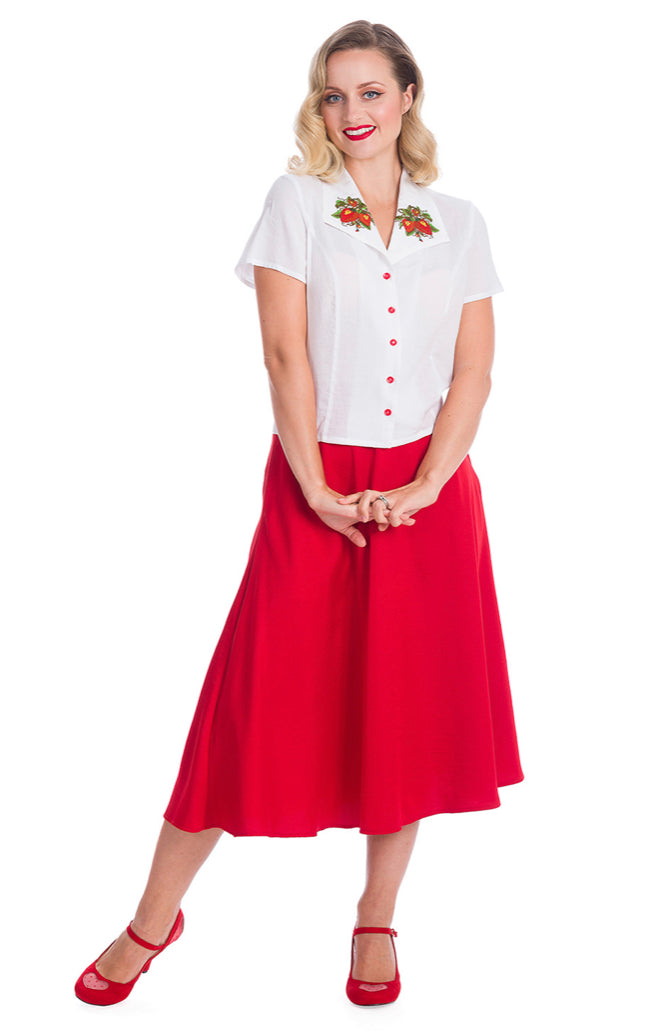 Blonde woman standing with her hands together in front of her. She is wearing a crisp white short sleeved blouse with red buttons and a wide 40s style collar with embroidered strawberries. 