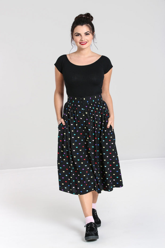 Smiling woman standing wearing black shoes, light pink socks, a black mid length skirt with multicoloured hearts all over and front black button detailing and a plain black top.
